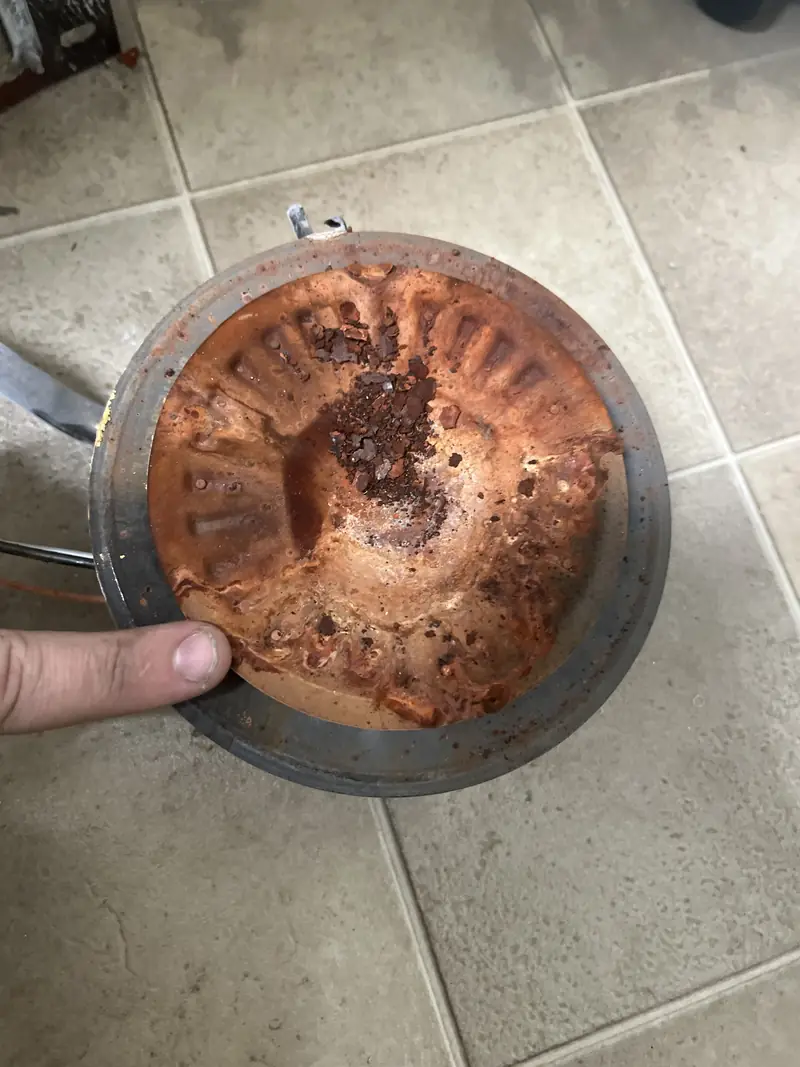 A heavily rusted round plate from the bottom of a water heater, held over a tiled floor