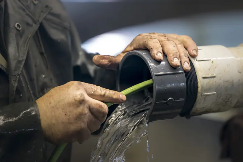 A plumber's hands holding a flexible hose inside a large pipe fitting while water pours out